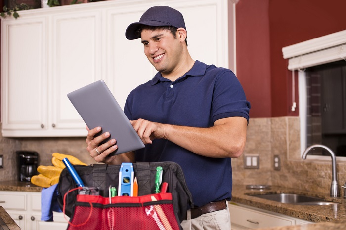 Hispanic repairman or blue collar/service industry worker makes service/house call inside customer's home kitchen. He has his tool box filled with work tools on counter as he uses his digital tablet.  Inspector, exterminator, electrician, plumber, repairmen.  He wears a navy blue uniform.